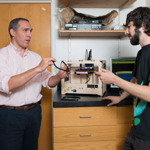 Sydney Cash, MD, PhD, holds a prototype of a device his lab is designing. Behind him, the 3D printing machine they use to create their designs.
